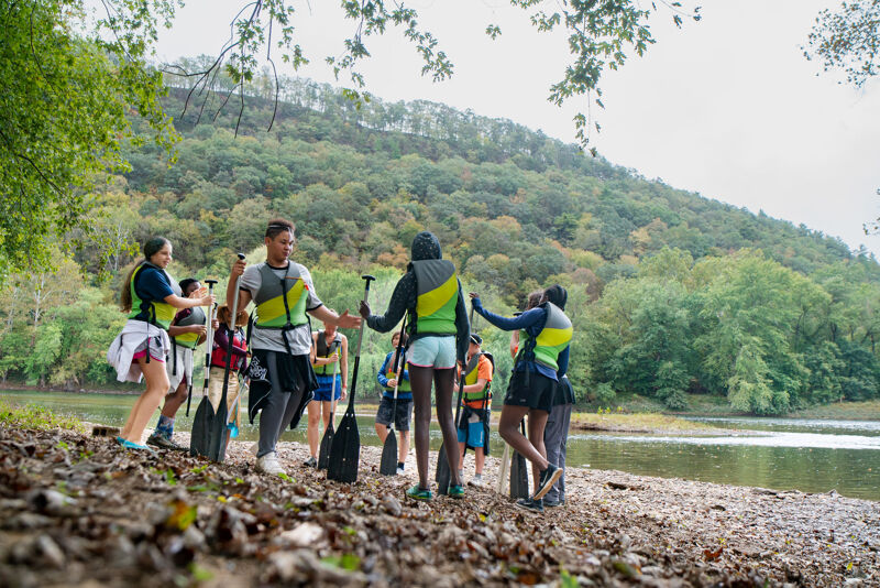A group of people wearing life jackets and holding paddles are gathered on a rocky shore next to a body of water. They appear to be preparing for or returning from a kayaking or canoeing trip. The background features a lush, green hillside and trees, suggesting a natural, outdoor setting.